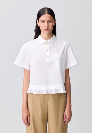 Blouse blanche à manches courtes avec un col, présentant une patte de boutonnage à l'avant avec des boutons dorés et un ourlet plissé. Une poche sur la poitrine est incluse.