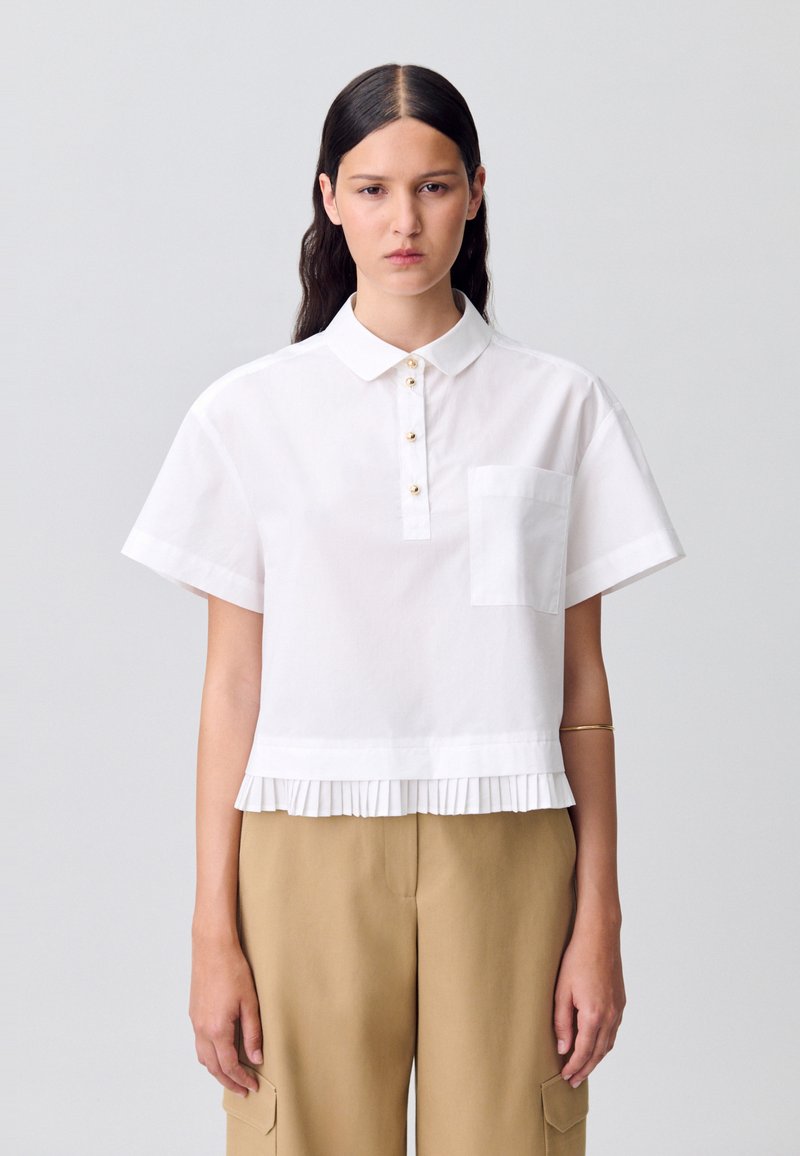 Blusa blanca de manga corta con cuello, que presenta una tapeta delantera con botones dorados y un dobladillo plisado. Incluye un bolsillo en el pecho.