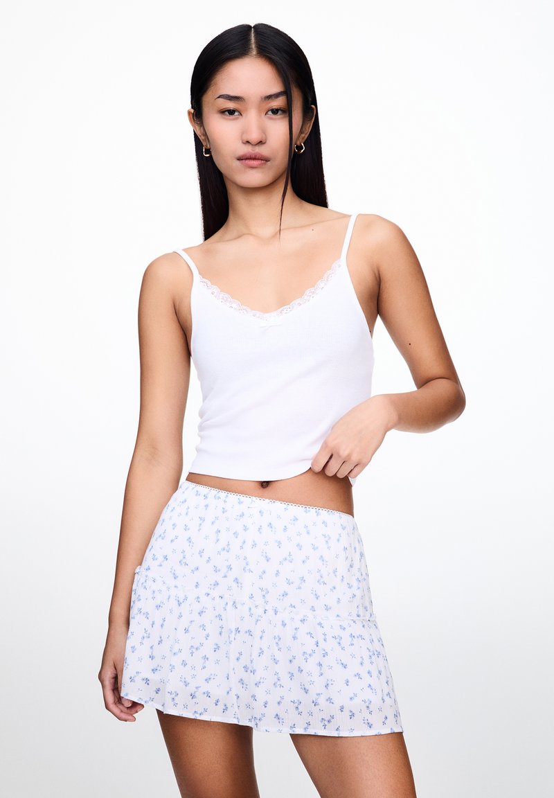 Weißes Tanktop mit Spitzendetails und einem hellblauen Blümchenrock mit rüschierten Stufen. Glatte Textur und taillierte Passform.