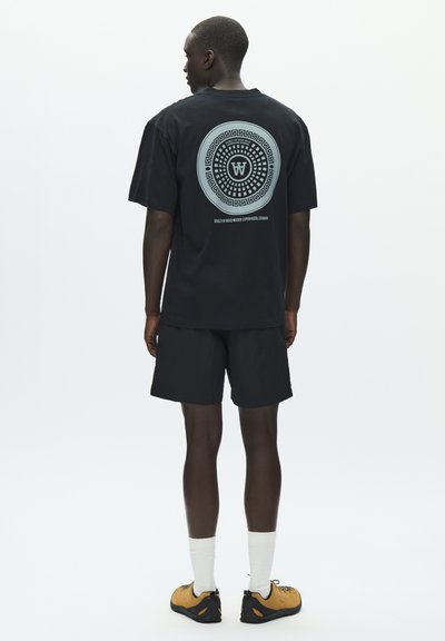 Μαύρο βαμβακερό t-shirt με κυκλικό γραφικό σχέδιο στην πλάτη, σε συνδυασμό με μαύρο σορτς και κίτρινα-μαύρα παπούτσια.