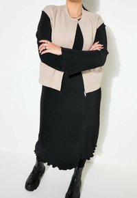 Beżowa kamizelka bez rękawów na czarnej prążkowanej sukience z falistym dołem; zestawiona z czarnymi botkami za kostkę. Obecne srebrne pierścionki i naszyjnik z łańcuszka.