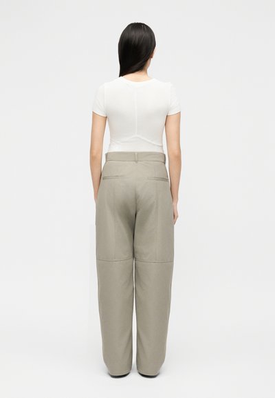 Femme aux longs cheveux noirs portant une chemise blanche ajustée à manches courtes et un pantalon beige ample taille haute, debout de dos sur un fond uni.