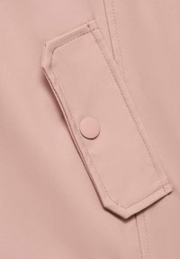 Detaliu al unei jachete roz deschis, cu o mare buzunar dotat cu închidere prin apăsare, textură netedă și cusături vizibile pe margini.