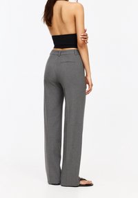 Vrouw in hoge, grijze broek, een zwart strapless top en zwarte sandalen, staand met de armen gebroken tegen een witte achtergrond.