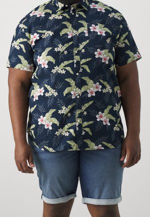 Hombre con una camisa azul marino de manga corta con botones, con patrones de hojas verdes y flores blanco-rosadas y pantalones cortos de mezclilla remangados.