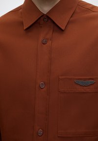 Chemise boutonnée de couleur rouille avec une poche sur la poitrine gauche comportant une épingle argentée avec le logo Aston Martin.