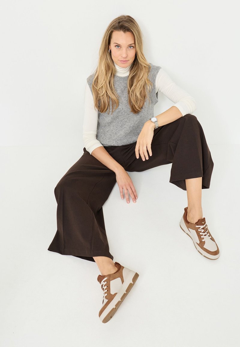 Grå ærmeløs sweater over en hvid langærmet skjorte, brune brede bukser og beige-brune sneakers med snørebånd og teksturerede detaljer.