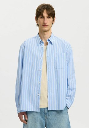 Ung mand iført løs blå- og hvidstribet skjorte med knapper over beige top og lyseblå jeans, stående foran en ensfarvet hvid baggrund.