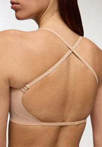 Reggiseno beige chiaro con design senza cuciture e sottili spalline incrociate. Presenta una chiusura a gancio e occhiello sul retro. Texture liscia.