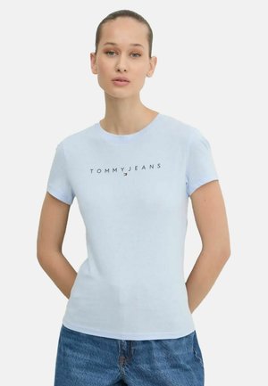 Donna che indossa una t-shirt Tommy Jeans azzurro chiaro e jeans blu, in piedi con le mani dietro la schiena davanti a uno sfondo uniforme.