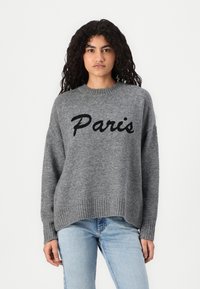 Femeie cu păr creț, purtând un pulover gri cu textul "Paris" în negru și blugi de un albastru deschis, pe un fundal alb simplu.