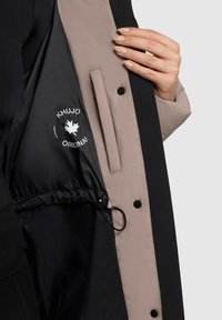 Îmbrăcăminte exterioară neagră și bej, cu interior ajustabil elastic și fermoar, având un logo tipărit și închideri cu capse. Textură moale și fină.