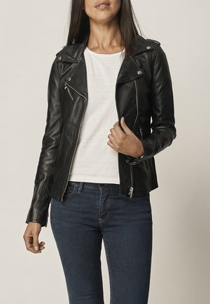 Femme portant une veste en cuir noire sur un t-shirt blanc et un jean bleu, tenant la veste près de la fermeture éclair sur un fond uni.