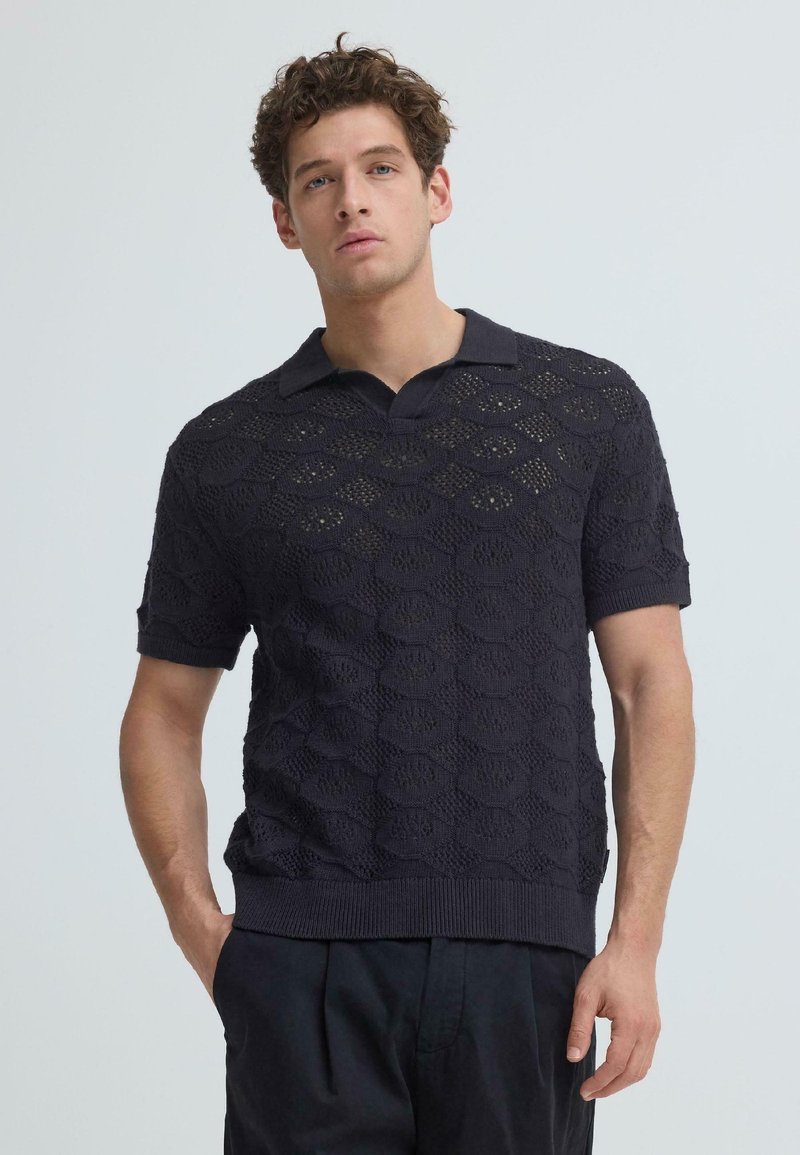 Νέος άνδρας με σγουρά μαλλιά, φοράει μπλε κοντομάνικο πόλο πλεκτό πουκάμισο και μαύρο παντελόνι, στέκεται μπροστά σε ένα απλό φόντο.