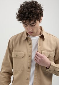 Beige Hemd mit Knöpfen, zwei Brusttaschen und sichtbaren Nähdetails. Über einem weißen T-Shirt getragen, mit Kragen und langen Ärmeln.
