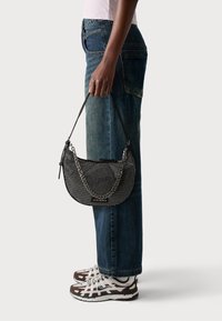 Black mesh handbag with a chain strap, featuring a half-moon shape and zip closure. The model wears blue jeans and athletic shoes.