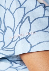 Světle modré bavlněné tričko s tmavomodrým květinovým vzorem. Logo G-STAR RAW v růžové barvě, umístěné na dolním levém rukávu.