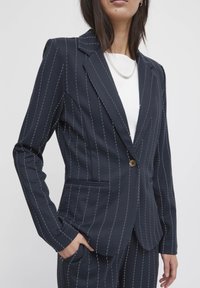 Marineblauer Nadelstreifen-Blazer mit einem einzigen Knopf, Reverskragen und strukturiertem Schnitt. Verfügt über seitliche Taschen und kontrastierende Nähte.