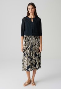 Cardigan nero con maniche a tre quarti, chiusura a bottoni, abbinato a una gonna fluida con un motivo di foglie beige su uno sfondo scuro.