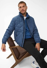 Blå quiltet jakke med høj krave, lynlås-lukning og sidelommer. Blevet parret med en blå sweater og sorte bukser, siddende på en træstol.