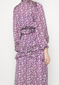 Robe à motifs floraux violets avec une taille cintrée, un ourlet froncé et des manches longues. Fabriquée en tissu léger avec une texture légèrement transparente.