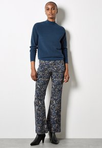 Granatowy sweter ribbed z wysokim kołnierzem, połączony z rozszerzanymi spodniami w wzór w odcieniach niebieskiego i złota, noszony z czarnymi botkami za kostkę.