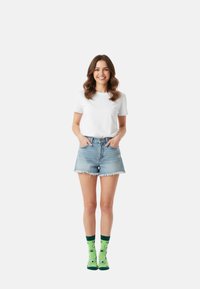 Grüne Socken mit Avocado-Muster, Jeans-Shorts mit fransigem Saum und ein schlichtes weißes T-Shirt. Das Model steht mit den Händen in den Taschen vor einem weißen Hintergrund.