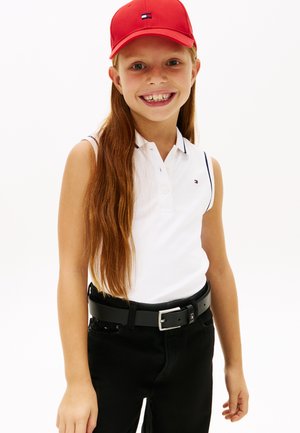 Usmievavá dievčina s dlhými červenými vlasmi, ktorá nosí červenú šiltovku, biele bezrukávové polo tričko, čierne nohavice a čierny opasok na bielom pozadí.