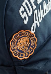 Zaino in tessuto navy con lettering ricamato bianco e un patch circolare arancione con un design di tigre e il testo "STAT ATHLETIC CHAMPIONSHIP."