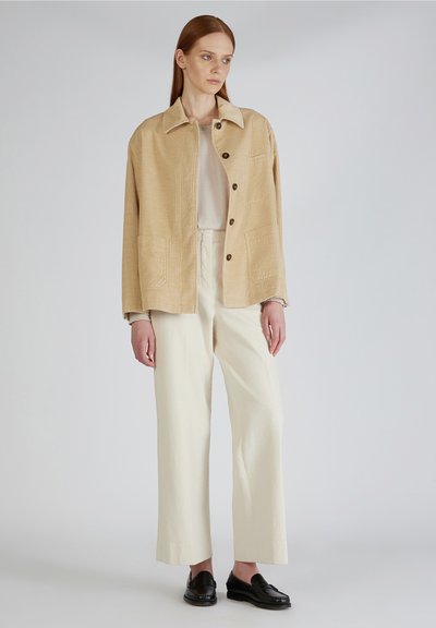 Beige Cord-Jacke mit Kragen, Knopfverschluss und zwei Fronttaschen. Kombiniert mit creamfarbenen, weit geschnittenen Hosen und schwarzen Slippern.