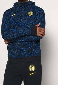 Sweat à capuche noir avec motif abstrait bleu, comprenant une poche avant, une capuche avec cordon de serrage, et un logo Nike jaune sur la poitrine. Poignets et ourlet noirs.