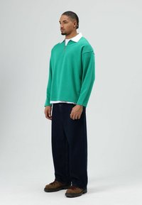 Groen sweatshirt met een witte kraag en ritssluiting, gecombineerd met donkerblauwe loszittende jeans en bruine veterlaarzen. Eenvoudig, casual ontwerp.