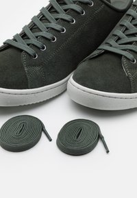 Grüne Wildledersneaker mit strukturierten Oberflächen, weißen Gummisohlen und passenden Schnürsenkeln. Zwei zusätzlich grüne Schnürsenkel neben den Schuhen.