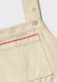 Bretelle de salopette en tissu beige avec deux boutons beiges et une poche frontale ornée d'une couture en zigzag rouge le long du bord supérieur.