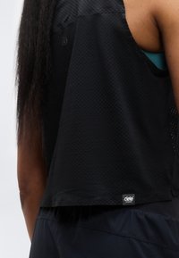 Haut de sport noir sans manches avec un design en maille texturée, doté d'une étiquette logo en bas. L'arrière est légèrement plus long que le devant.