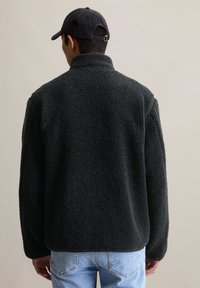 Veste en polaire noire avec un col haut, texture douce et manches longues, portée avec un jean bleu clair sur un fond neutre.