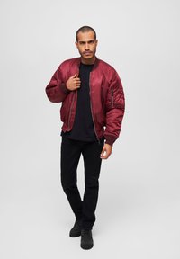 Veste bomber bordeaux avec des poignets côtelés, des poches zippées et une finition satinée, portée sur un t-shirt noir et un pantalon noir.
