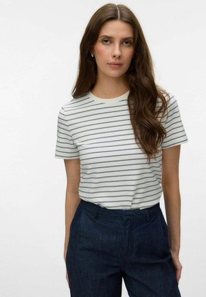 Kvinde med langt brunt hår iført en hvid t-shirt med korte ærmer og tynde vandrette striber samt højtaljede mørkeblå bukser mod en ensfarvet baggrund.