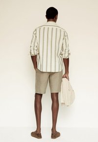 Gestreept shirt met lange mouwen in off-white en beige, lichtbeige korte broek, en bruine instapschoenen. Houdt een jas in lichte tint vast.