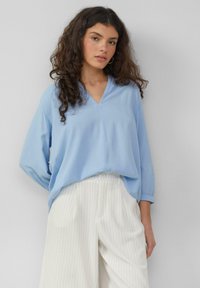Lichtblauwe blouse met lange mouwen en een V-hals, gemaakt van een zachte stof, gecombineerd met witte broeken met streepjes en een knoopsluiting.