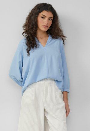 Lichtblauwe blouse met lange mouwen en een V-hals, gemaakt van een zachte stof, gecombineerd met witte broeken met streepjes en een knoopsluiting.