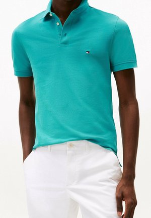 Mand iført turkis kortærmet polo-shirt med lille logo, kombineret med hvide bukser, hænderne i lommerne, foran en ensfarvet baggrund.