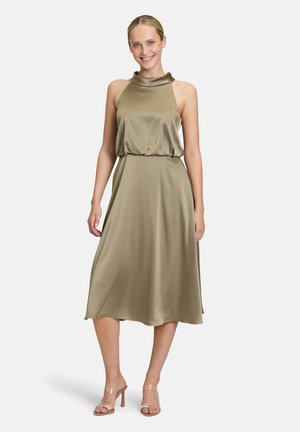 Olivgrünes Midi-Kleid mit Halterneck, gerafftem Taillenbund und seidiger Textur. Kombiniert mit transparenten Riemchensandalen.