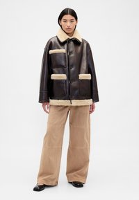 Giacca di pelle marrone con collo peloso crema e tasche, abbinata a pantaloni larghi color cammello e scarpe slip-on nere.