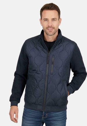 Marineblauw gewatteerd vest met hoge kraag, ritssluiting aan de voorkant en zijzakken. Gemaakt van gladde stof, met contrasterende texturen en accenten.