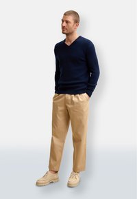 Marinblå V-ringad tröja, beige byxor och ljusbruna skor. Tröjan har ribbade manschetter, byxorna är raka med en slät textur.