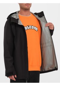 Czarna kurtka z kapturem i zapięciem na zamek, z jasnoszarym wewnętrznym podszyciem. Noszona na pomarańczowej bluzie z nadrukiem graficznym.