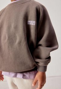 Kind trägt braunes Sweatshirt mit dem Aufdruck "AMERICAN VINTAGE" auf der Brust, darüber ein fliederfarbenes Hemd und helle Hosen.