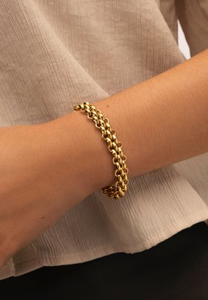 Bracelet en double chaîne dorée porté au poignet sur un fond de tissu beige texturé et de pantalons sombres.