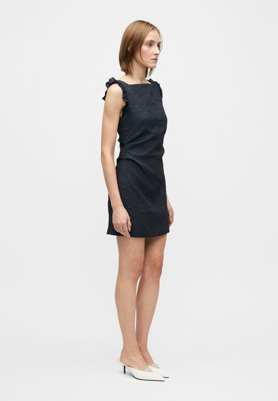 Rochie bleumarin din țesătură texturată, cu accente plisate pe umeri și croială mulată, purtată cu mule cu toc albe.